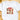 A white short-sleeve crewneck t-shirt laid flat on a light wooden surface, featuring a festive Christmas collage graphic on the chest with a red bow, snowman, nutcracker, Santa, reindeer, and red truck.