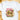 A white short-sleeve crewneck t-shirt laid flat on a light wooden surface, featuring a colorful graphic of a green cat wearing a pink leopard-print Santa hat and pink sunglasses, with the words 'MERRY BRIGHT' in pink and olive green.