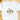 A white short-sleeve crewneck t-shirt laid flat on a light wooden surface, featuring a watercolor-style Christmas church graphic with the text 'O Come Let Us Adore Him' and small decorative hearts.