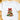 A white short-sleeve crewneck t-shirt laid flat on a light wooden surface, featuring a watercolor-style Christmas tree graphic with a large red bow and green and red tree layers.
