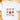 A white short-sleeve crewneck t-shirt laid flat on a light wooden surface, featuring a watercolor-style Christmas graphic print with a mug, bows, deer, Santa, snowman, and a Christmas tree.