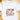 A white short-sleeve crewneck t-shirt laid flat on a light wooden surface, featuring a colorful 'Merry Christ Mas' graphic with illustrated Christmas trees, holly, and gold stars.