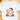 A white short-sleeve crewneck t-shirt laid flat on a light wooden surface, featuring a colorful watercolor graphic of a cute ghost wearing a red Santa hat and wrapping multicolor Christmas lights, with wrapped presents at its feet.