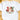A white short-sleeve crewneck t-shirt laid flat on a light wood surface, featuring a festive Christmas graphic with the word 'JOY' in red and green letters, a Santa Claus face, and the phrase 'To the world' in green script.